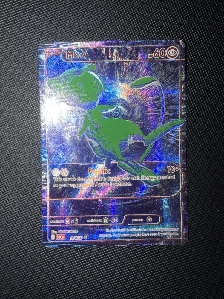 Carte Mew B/RGB du set 30C sortie de sa protection, posée à plat sur un tapis noir texturé, fond bleu-violet holographique et Mew vert, attaque Psychic 10+ dégâts, illustration YOSHIROTTEN